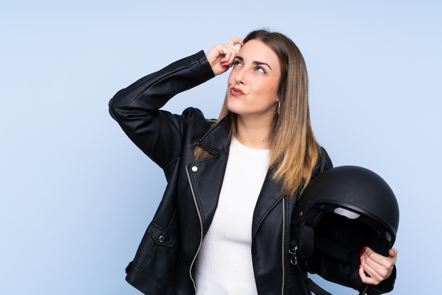 Jovem mulher loira com um capacete de moto sobre parede azul isolada, tendo dúvidas e com a expressão do rosto confuso