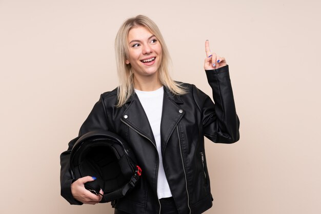 Jovem mulher loira com um capacete de moto sobre parede isolada, com a intenção de realizar a solução enquanto levanta um dedo