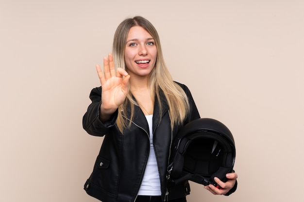 Jovem mulher loira com um capacete de moto sobre parede isolada, mostrando um sinal de ok com os dedos