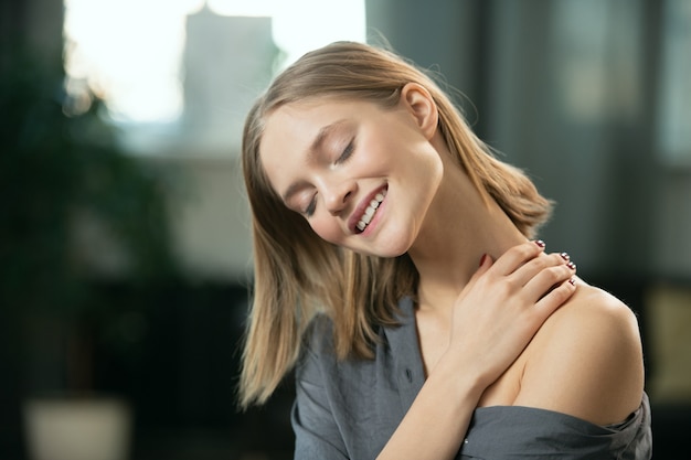 Jovem mulher loira com um sorriso cheio de dentes, aplicando um produto cosmético natural feito à mão no pescoço e no corpo e desfrutando de manhã