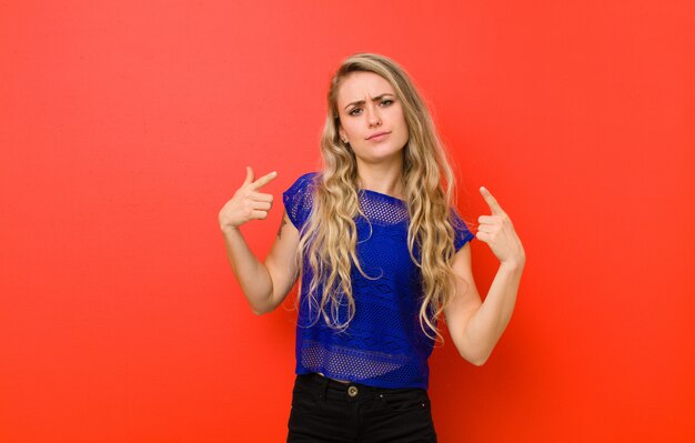 Jovem mulher loira com uma atitude ruim, olhando orgulhoso e agressivo, apontando para cima ou fazendo graça sinal com as mãos contra a parede vermelha