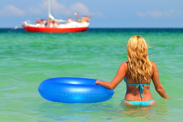 Jovem mulher loira de biquíni azul em pé para trás na água do mar parada com círculo de natação e olhando para o navio no horizonte num dia ensolarado de verão. Conceito de felicidade, férias e liberdade