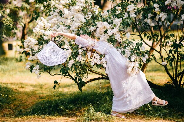 Jovem mulher loira de vestido branco, posando em arbustos lilás