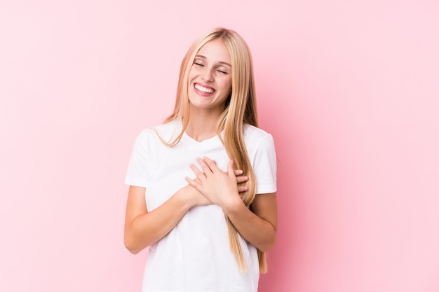 Jovem mulher loira em fundo rosa rindo mantendo as mãos no coração