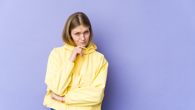 Jovem mulher loira isolada em um fundo roxo, olhando de soslaio com expressão duvidosa e cética.