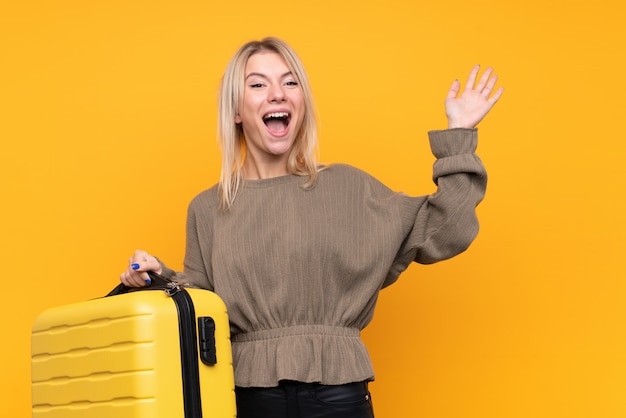 Jovem mulher loira isolado parede amarela em férias com mala de viagem e surpreso