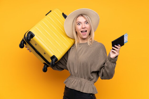 Jovem mulher loira isolado parede amarela em férias com mala e passaporte
