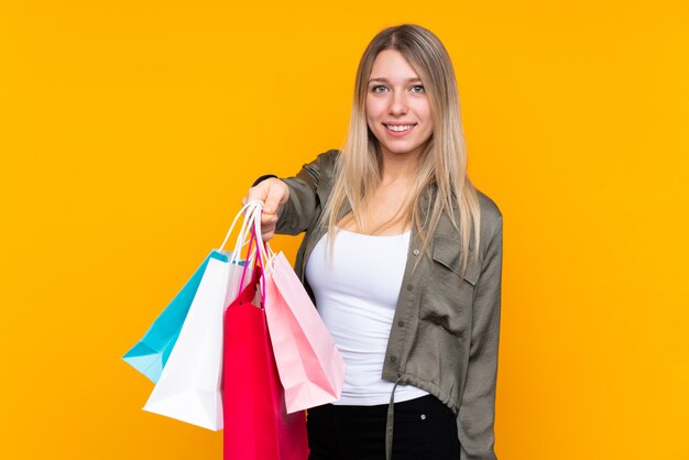 Jovem mulher loira isolado parede amarela segurando sacolas de compras e dar a alguém