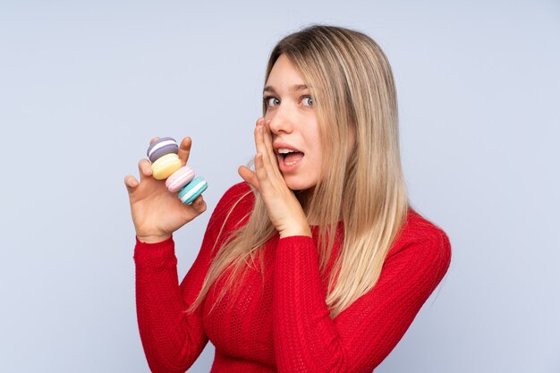 Jovem mulher loira isolado parede azul segurando macarons franceses coloridos e sussurrando algo