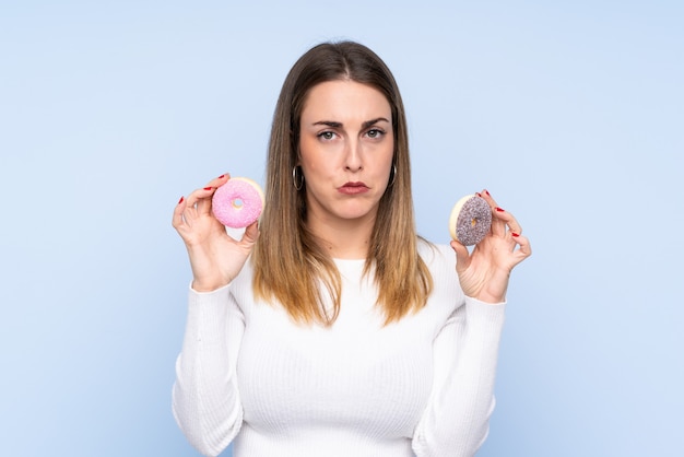 Jovem mulher loira isolado parede azul segurando rosquinhas com expressão triste