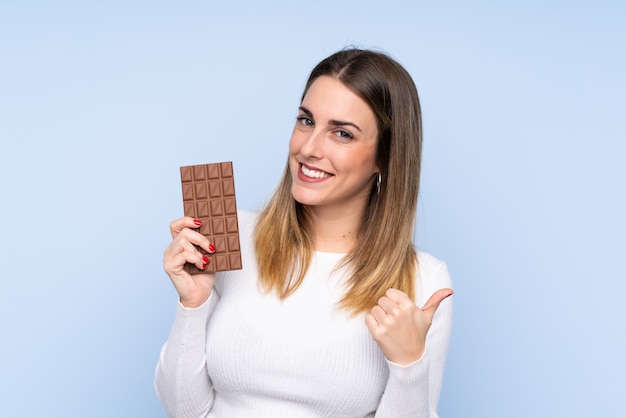 Jovem mulher loira isolado parede azul tomando uma tablete de chocolate e com o polegar