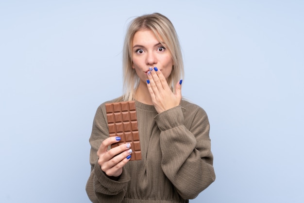 Foto jovem mulher loira isolado parede azul tomando uma tablete de chocolate e surpreendeu