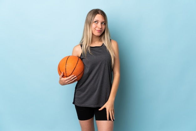 Jovem mulher loira jogando basquete isolado na parede azul de pé e olhando para o lado
