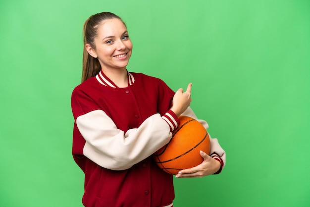 Jovem mulher loira jogando basquete sobre fundo croma isolado apontando para trás