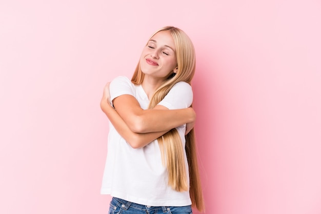 Jovem mulher loira na parede rosa abraços, sorrindo despreocupado e feliz.