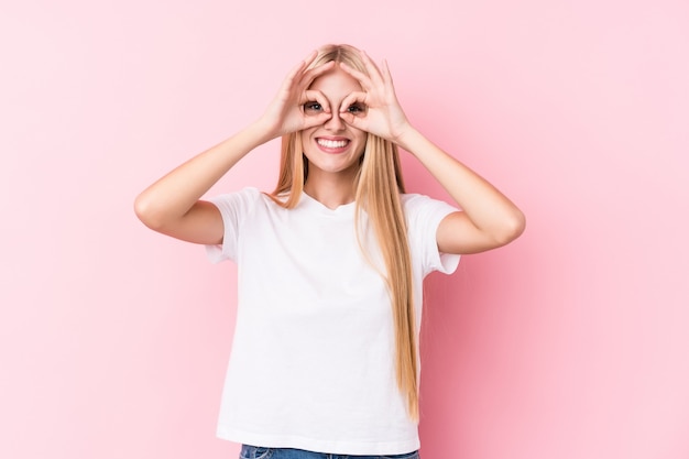 Jovem mulher loira na parede rosa mostrando sinal bem sobre os olhos