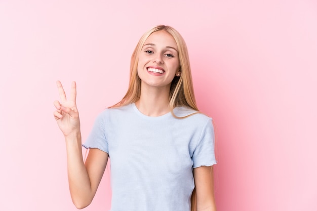 Jovem mulher loira na parede rosa mostrando sinal de vitória e sorrindo amplamente.