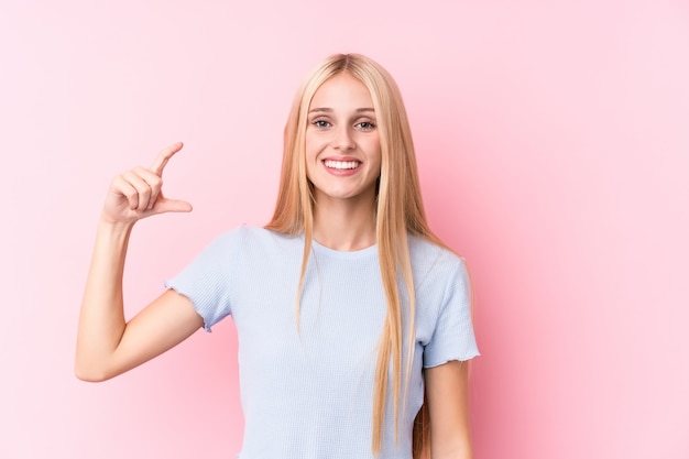 Jovem mulher loira no fundo rosa segurando algo pouco com o dedo indicador, sorrindo e confiante.