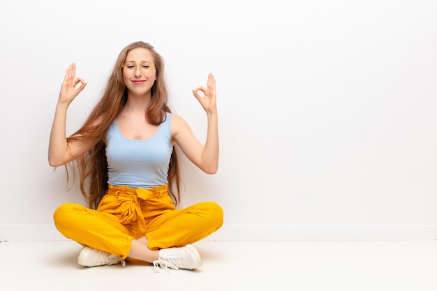 Foto jovem mulher loira olhando concentrada e meditando, sentindo-se satisfeita e relaxada, pensando ou fazendo uma escolha sentada no chão