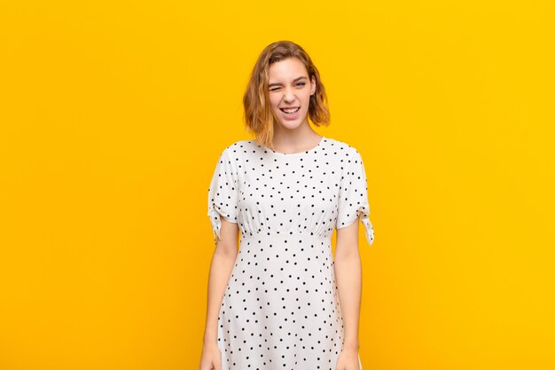 Jovem mulher loira olhando feliz e amigável, sorrindo e piscando um olho para você com uma atitude positiva contra a parede de cor lisa