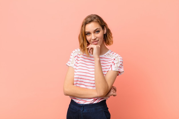 Jovem mulher loira olhando feliz e sorrindo com a mão no queixo, pensando ou fazendo uma pergunta, comparando opções contra parede plana de cor