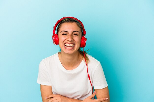 Jovem mulher loira ouvindo música em fones de ouvido isolados em um fundo azul, rindo e se divertindo.