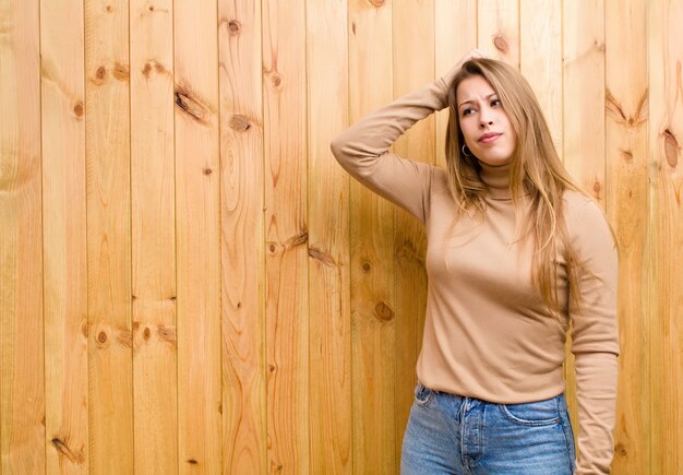 Jovem mulher loira se sentindo confusa e confusa, coçando a cabeça e olhando para o lado