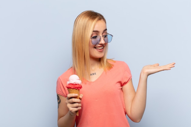 Jovem mulher loira se sentindo feliz e sorrindo casualmente, olhando para um objeto ou conceito seguro na mão ao lado