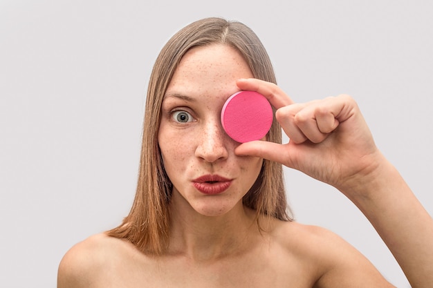 Jovem mulher loira segura esponja rosa e cobre os olhos com ele. Ela está espantada e maravilhada. Modelo parece.