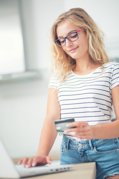 Jovem mulher loira segurando o cartão de crédito e usando o computador portátil na cozinha de casa.