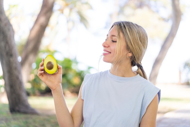 Jovem mulher loira segurando um abacate ao ar livre com expressão feliz