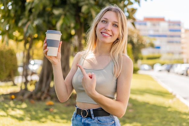 Jovem mulher loira segurando um café para viagem ao ar livre e apontando-o