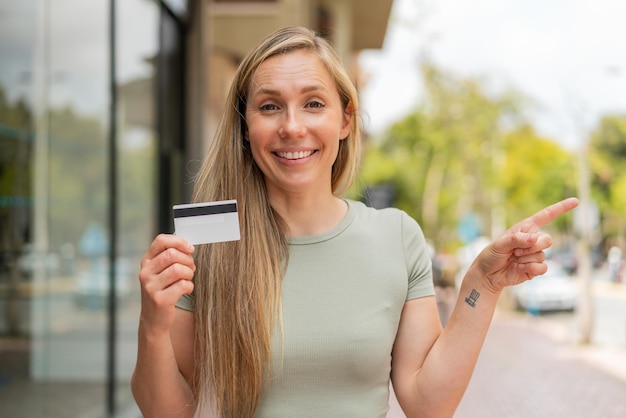 Jovem mulher loira segurando um cartão de crédito ao ar livre surpreso e apontando o dedo para o lado