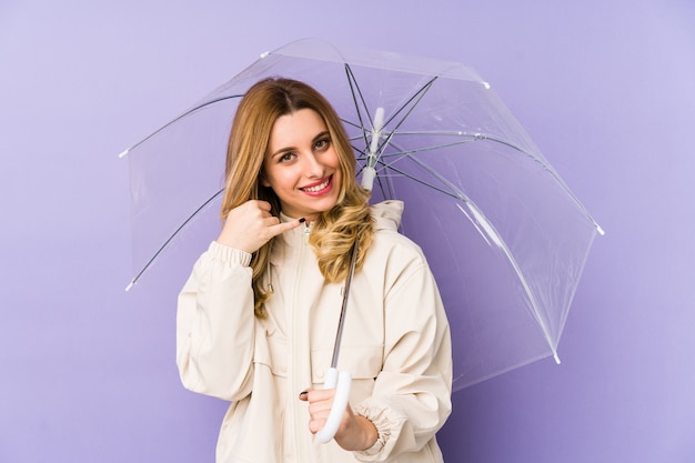 Jovem mulher loira segurando um guarda-chuva e mostrando um gesto de chamada de celular com os dedos.