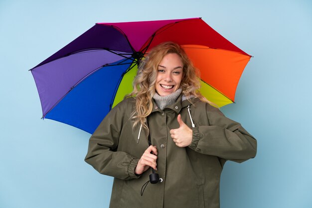 Jovem mulher loira segurando um guarda-chuva isolada na parede azul com polegares para cima, porque algo de bom aconteceu