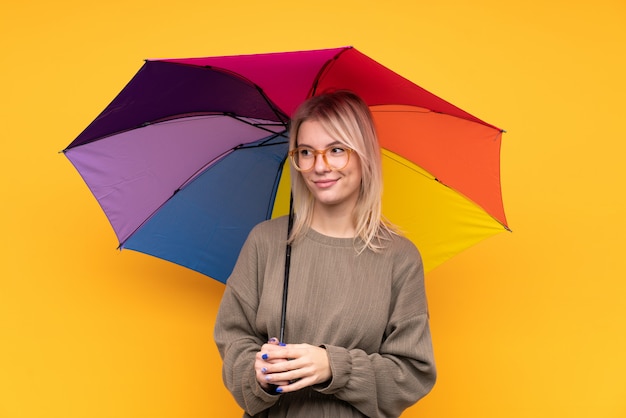 Jovem mulher loira segurando um guarda-chuva sobre parede amarela isolada em pé e olhando para o lado