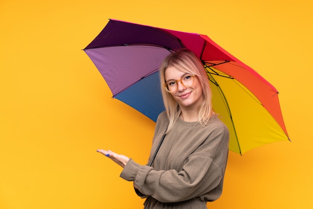 Jovem mulher loira segurando um guarda-chuva sobre parede amarela isolada, estendendo as mãos para o lado para convidar para vir