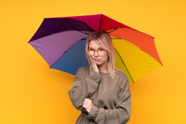 Jovem mulher loira segurando um guarda-chuva sobre parede amarela isolada infeliz e frustrada