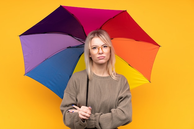 Jovem mulher loira segurando um guarda-chuva sobre parede amarela isolada, mantendo os braços cruzados