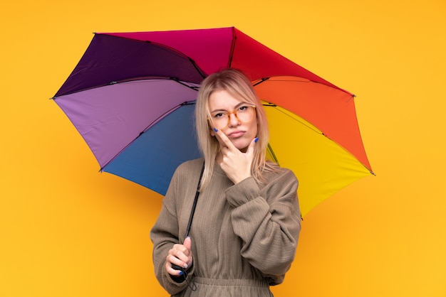 Jovem mulher loira segurando um guarda-chuva sobre parede amarela isolada, pensando em uma idéia