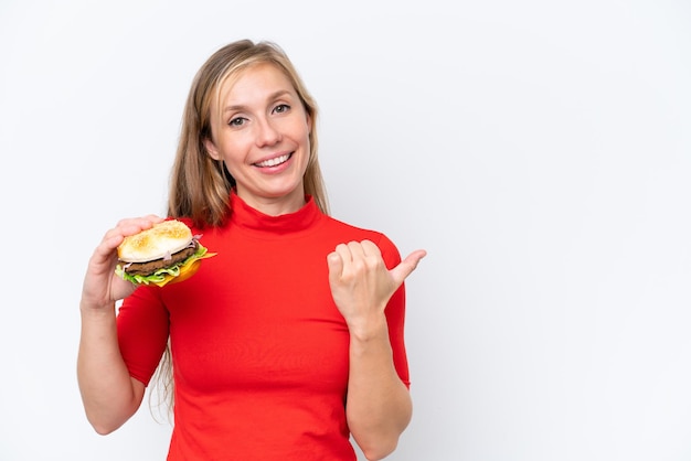 Jovem mulher loira segurando um hambúrguer isolado no fundo branco, apontando para o lado para apresentar um produto
