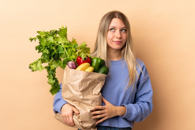 Jovem mulher loira segurando um saco cheio de legumes em pé e olhando para o lado