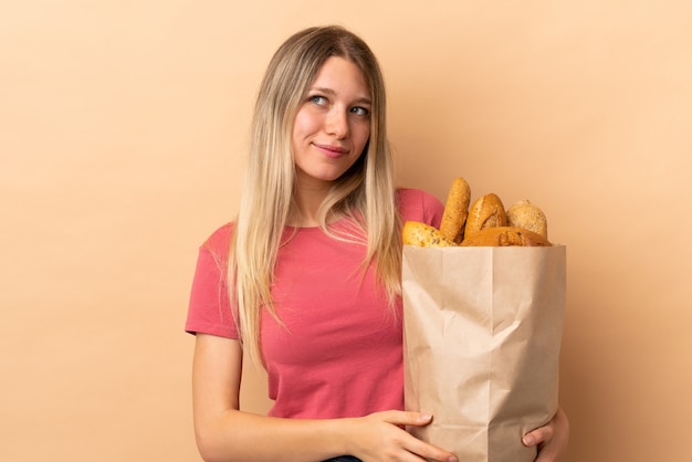 Jovem mulher loira segurando um saco cheio de pães isolados na parede bege de pé e olhando para o lado