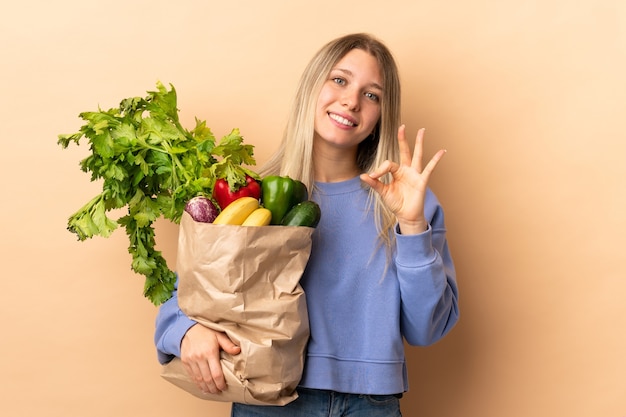 Jovem mulher loira segurando um saco cheio de vegetais isolado e mostrando um sinal de ok com os dedos