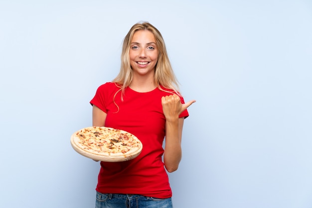 Foto jovem mulher loira segurando uma pizza sobre parede azul isolada, apontando para o lado para apresentar um produto