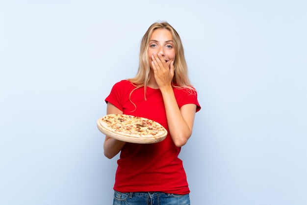 Jovem mulher loira segurando uma pizza sobre parede azul isolada com expressão facial de surpresa