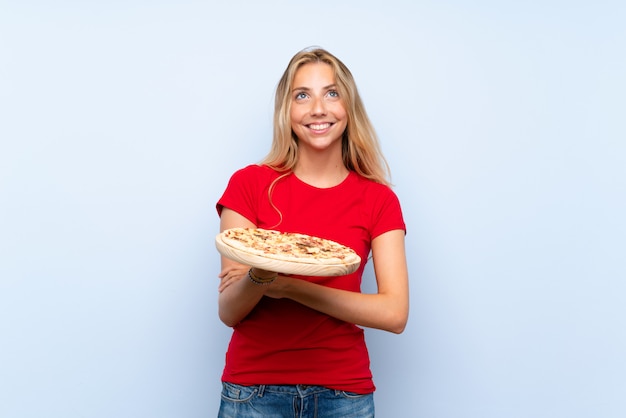 Foto jovem mulher loira segurando uma pizza sobre parede azul isolada, olhando para cima enquanto sorrindo