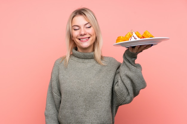 Jovem mulher loira segurando waffles sobre parede rosa isolada com expressão feliz