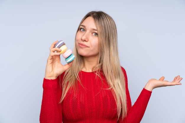 Jovem mulher loira sobre parede azul isolada segurando macarons franceses coloridos e fazendo gesto de dúvidas