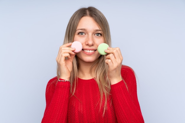 Jovem mulher loira sobre parede azul isolada segurando macarons franceses coloridos e sorrindo